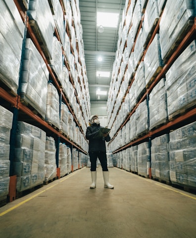 Warehouse aisle with tall shelves stacked with packages and a worker inspecting inventory, symbolizing logistics and supply chain management